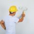 Builder man painting wall with roller in white t-shirt, helmet and looking busy , back view.